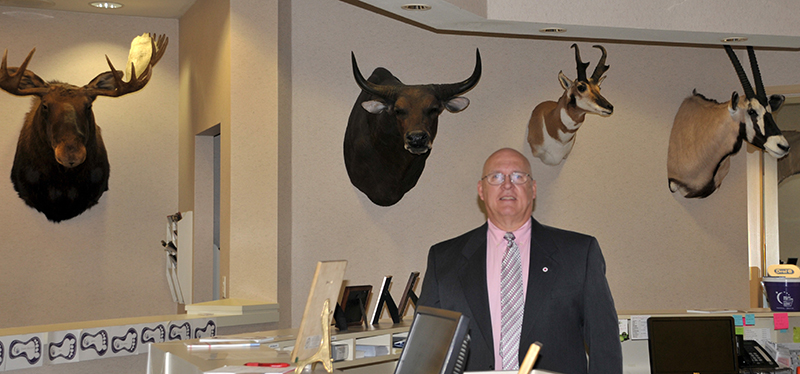Dr. Karl Clinard in his front office at Somerset with four wild game trophies on thewall behind him. From left are a Canadian moose; a Banteng wild cow from northern Australia; an Antelope from Wyoming; and a Gymsbock from Africa. There is a story with every one. For instance the Benteng is the most rare animal he has ever hunted. They were about a hundred miles from the nearest road, walking through crocodile infested waters where everything that can bite or sting waits for you. (Campbellsville University Photo by Linda Waggener)
